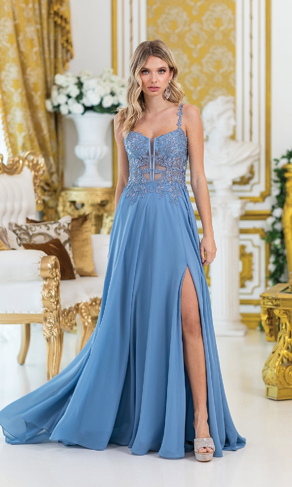 A woman in the Lace-Corset Long Prom Dress: Dancing Queen 4531, a blue embroidered lace gown with a high slit, stands in an ornate gold-accented room adorned with floral decor.